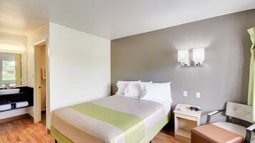 A hotel room with a bed, a bedside table with a lamp, a chair, and a bathroom with a sink and mirror.