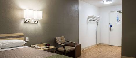 Habitación de hotel con una cama, una silla, un escritorio con teléfono y control remoto, una lámpara de pared, una puerta con perchero y suelo de madera.