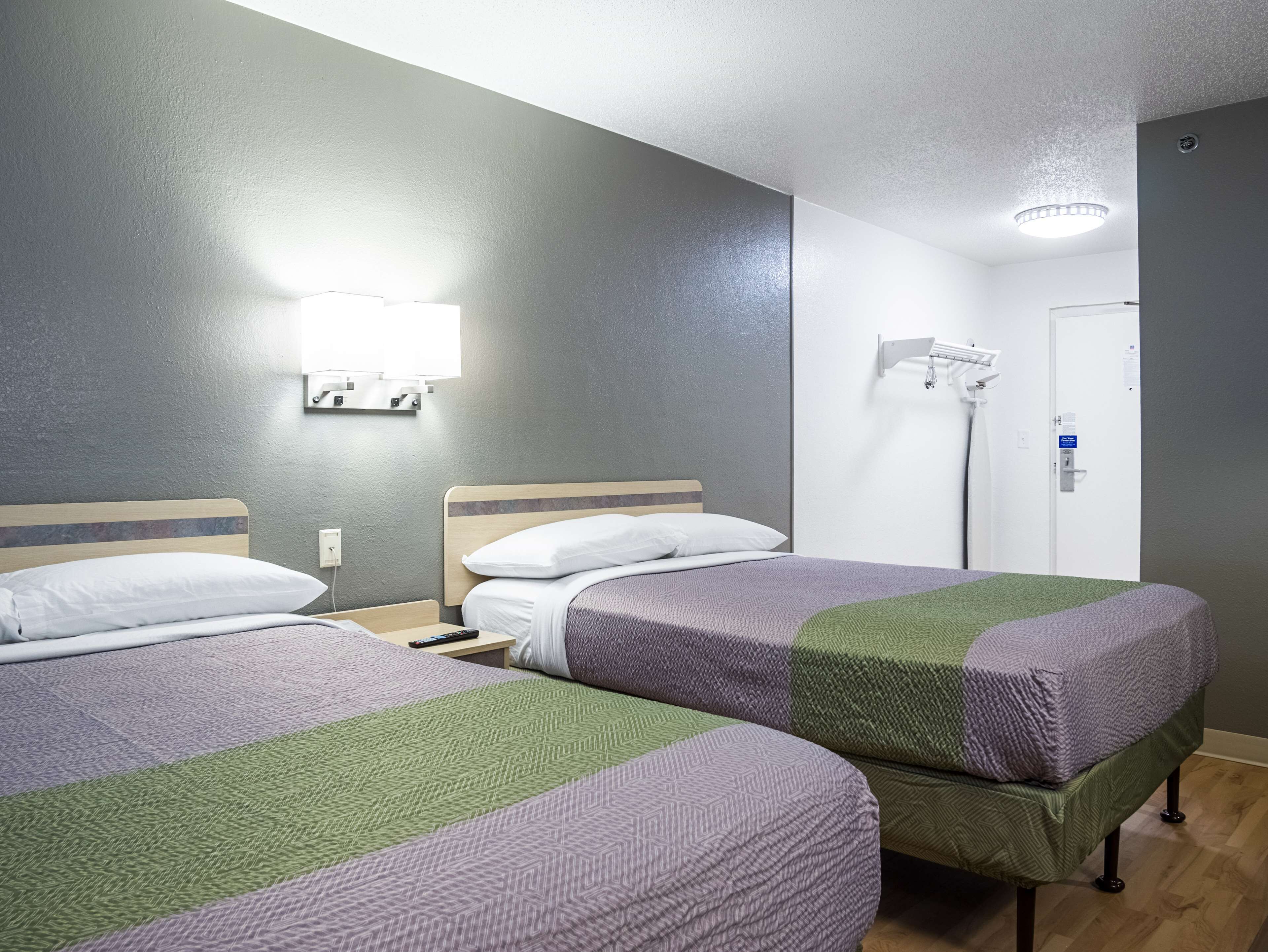A hotel room with two beds, a bedside table, a lamp, a mirror, and a door with a security camera.