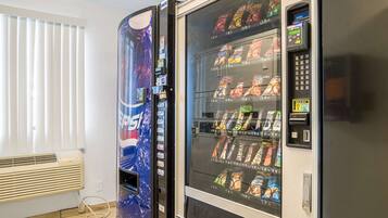 Two vending machines with snacks and drinks, a window with blinds, and a wall-mounted air conditioner.