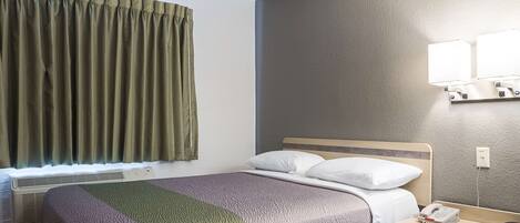 Una habitación de hotel con una cama, una mesita de noche con teléfono y control remoto, una ventana con cortinas y una lámpara fijada a la pared.