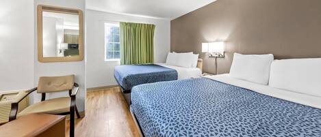 A hotel room with two beds, a chair, a desk, a mirror, a window with curtains, and a wall-mounted lamp.