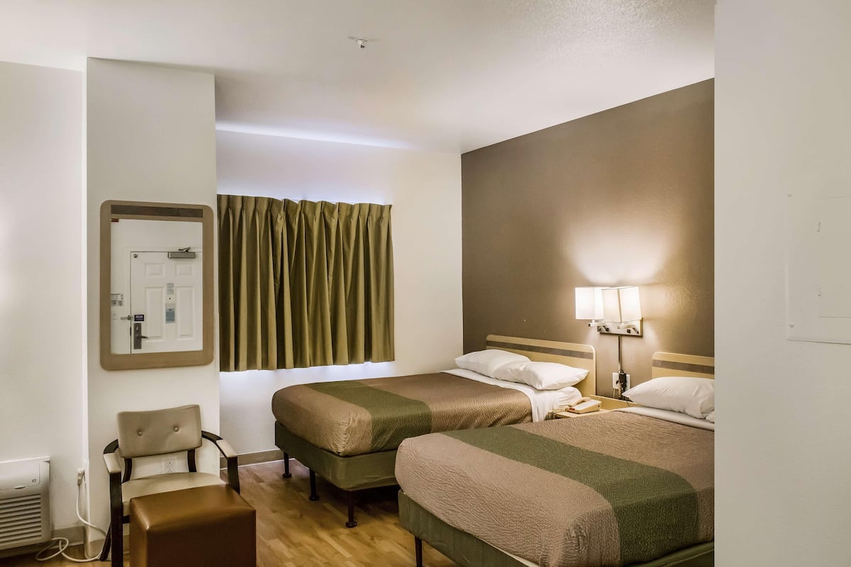 A hotel room with two beds, a chair, a desk, a lamp, and a window with curtains.