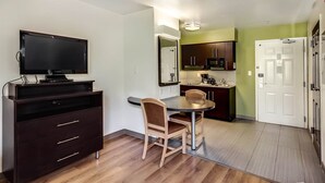 Een knus woongedeelte met een flatscreen televisie, een eettafel met twee stoelen, een keukenhoek met een magnetron en een witte deur.