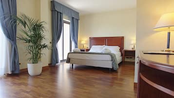 Camera d'albergo con pavimento in legno, un letto con lenzuola bianche, un comodino con una lampada e una pianta in vaso.