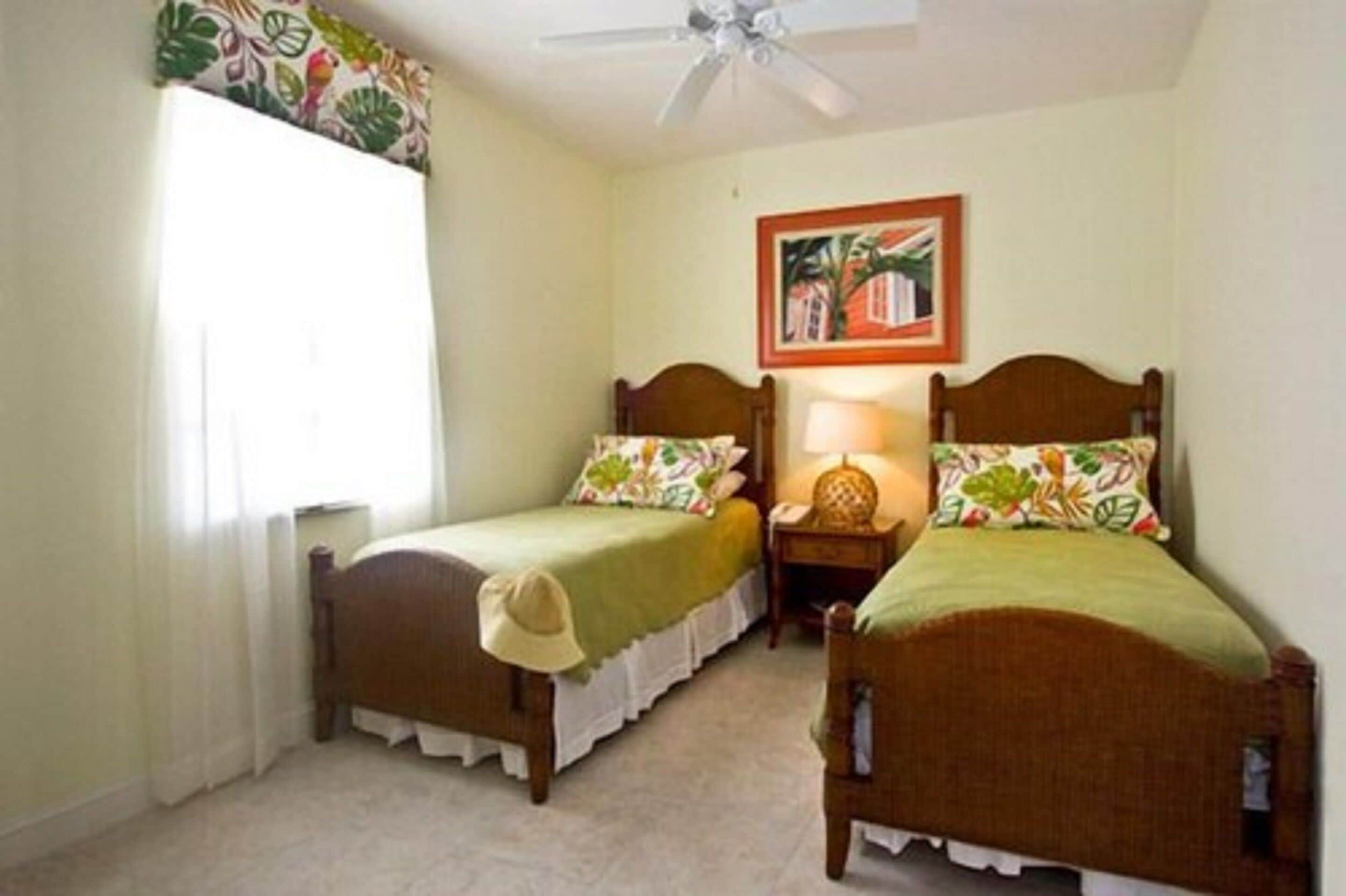 A room with two wooden beds, a ceiling fan, a window with curtains, and a framed picture on the wall.