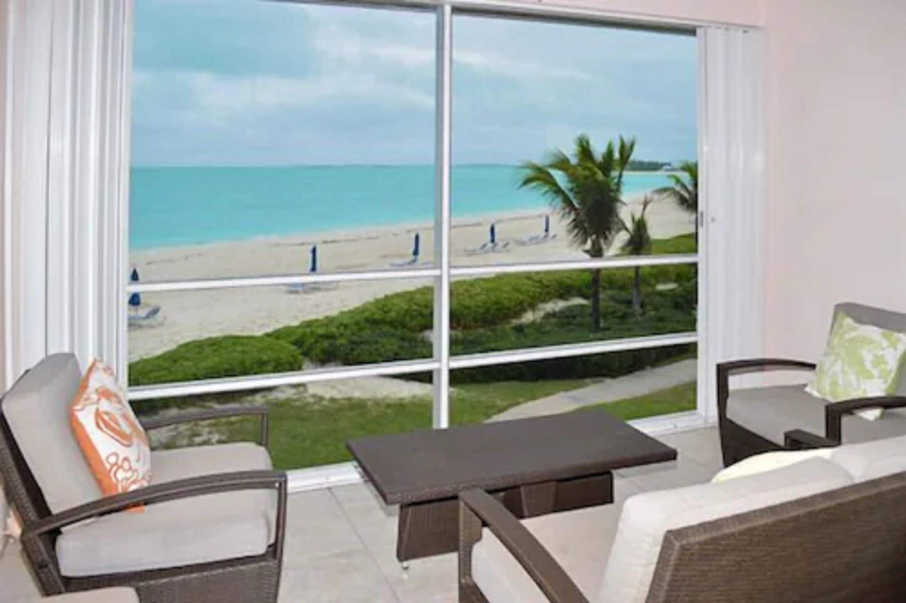 A balcony with a view of a beach, two chairs, and a small table.