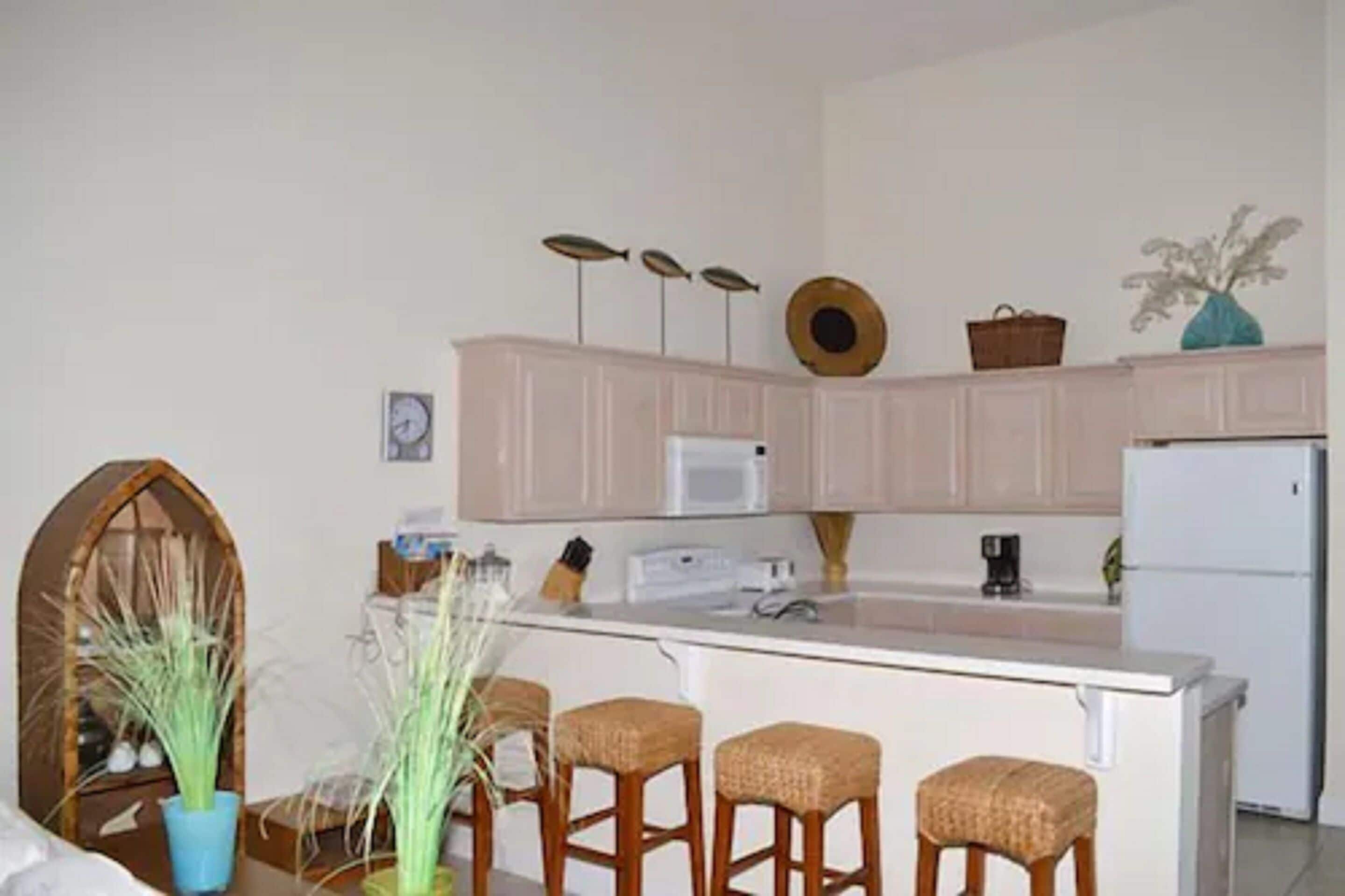 A kitchen with white cabinets, a white refrigerator, a coffee maker, and three wicker bar stools.