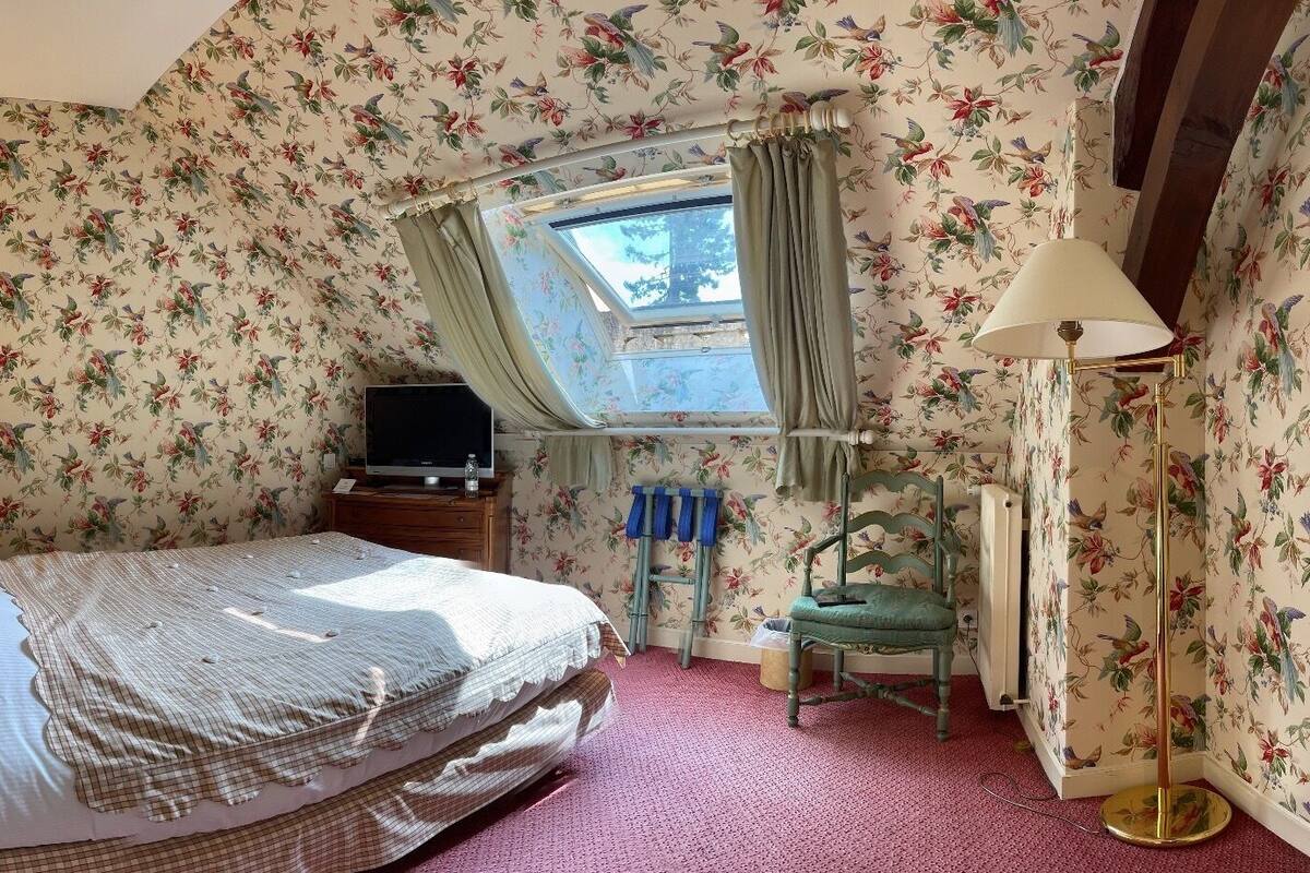 A bedroom with floral wallpaper, a bed with a checkered bedspread, a bedside table with a lamp, a chair, and a window with curtains.