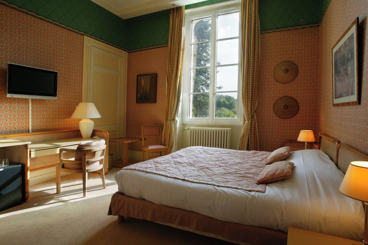 A bedroom with a bed, a desk, a chair, a television, and a window with curtains.