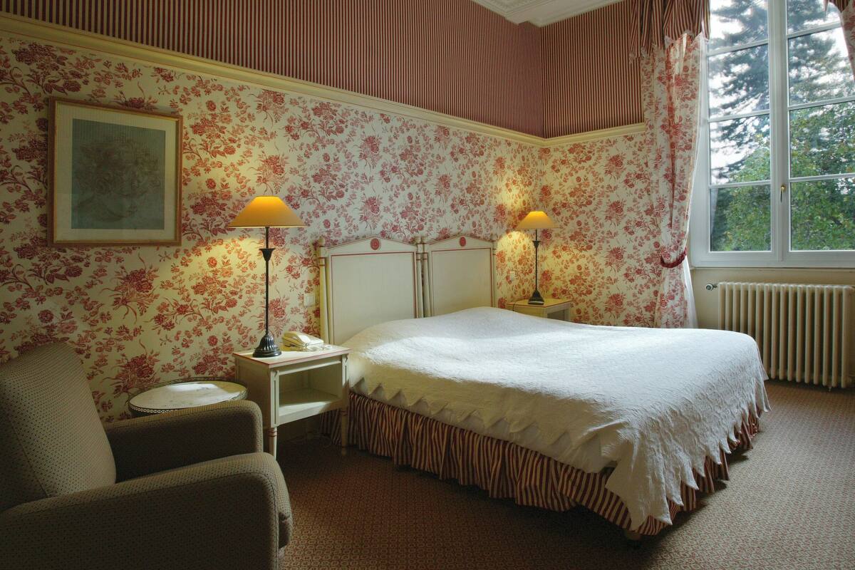 A bedroom with a bed, two bedside tables, a chair, a window, and floral wallpaper.