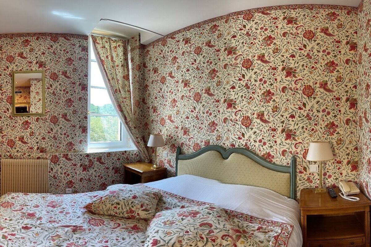 A bedroom with floral wallpaper, a bed with floral bedding, a nightstand with a lamp and phone, and a window with curtains.