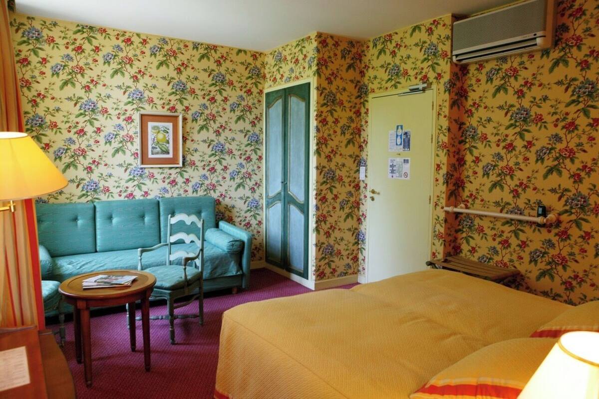 A room with floral wallpaper, a green sofa, a bed with a yellow bedspread, a small round table, and a lamp.