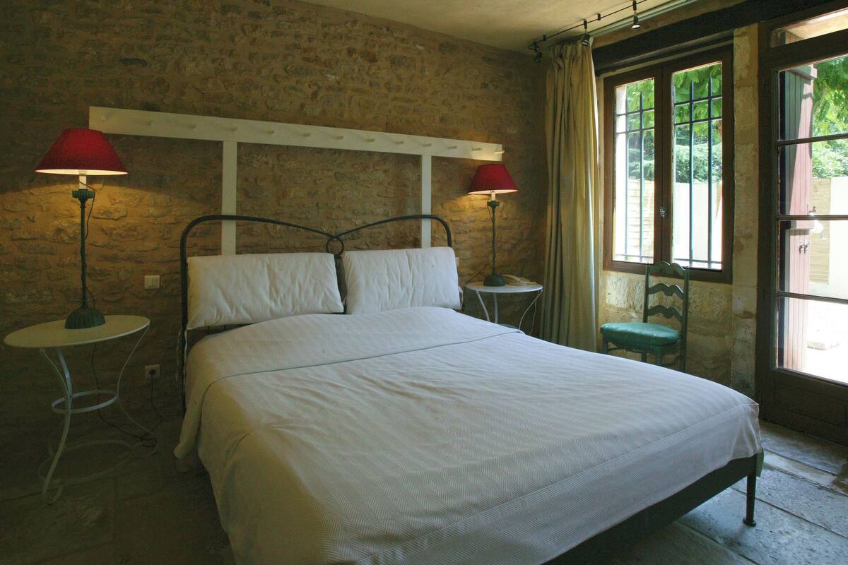 A bedroom with a bed, two bedside tables, a lamp on each table, a window with curtains, and a stone wall.