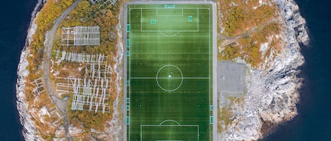 Eine Vogelperspektive zeigt ein Küstenanwesen mit großem Sportplatz, Parkbereich und umgebendem felsigem Gelände.
