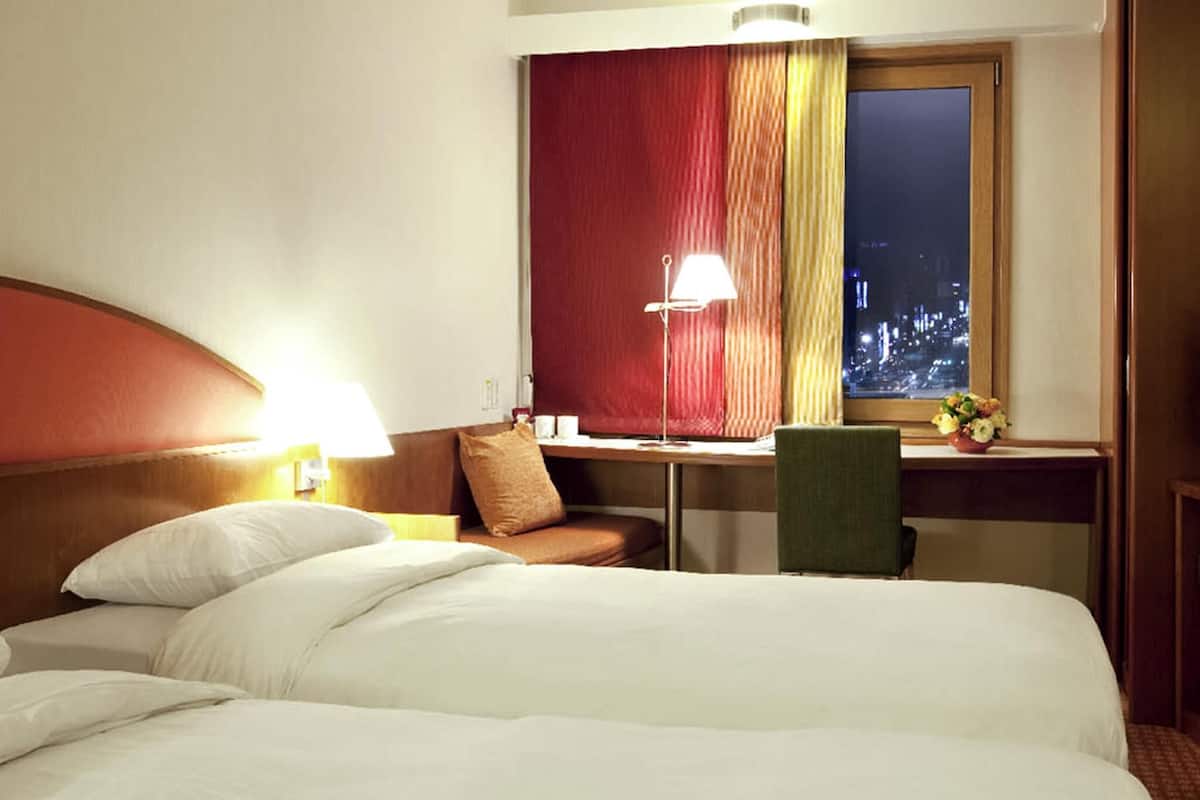 Une chambre d’hôtel avec un lit, un bureau, une chaise, une lampe et une fenêtre avec des rideaux.