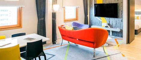 A hotel room with a dining area, a red sofa, a TV, and a bed.