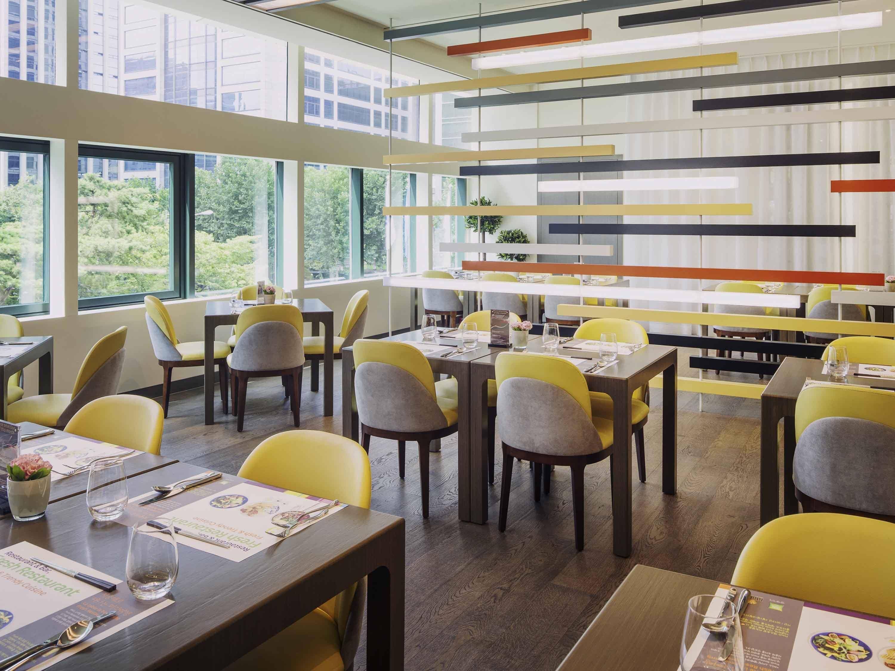 A modern restaurant interior with high ceilings, large windows, and yellow chairs.