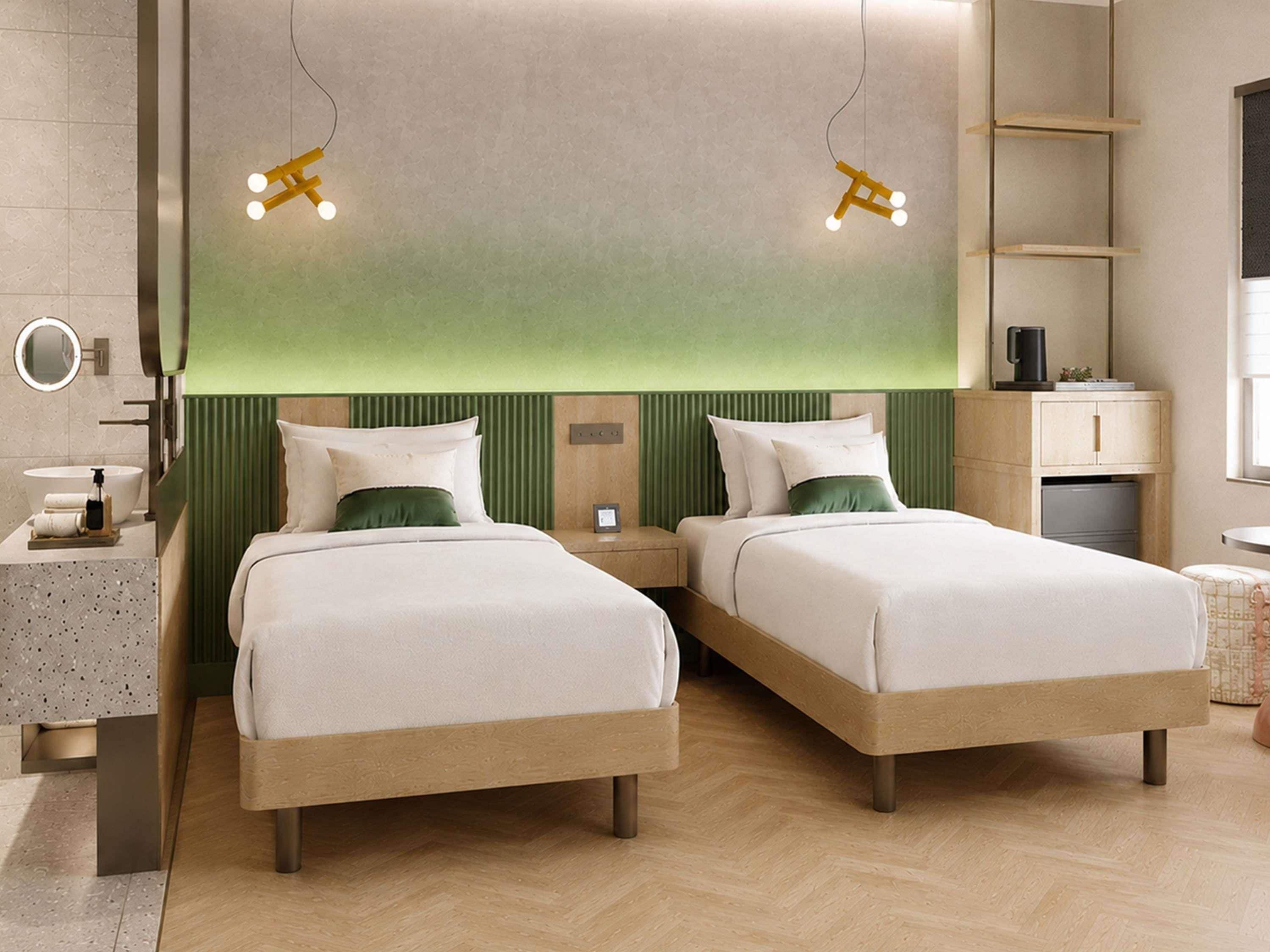 A hotel room with two beds, a bedside table, a mirror, a sink, and a green accent wall.