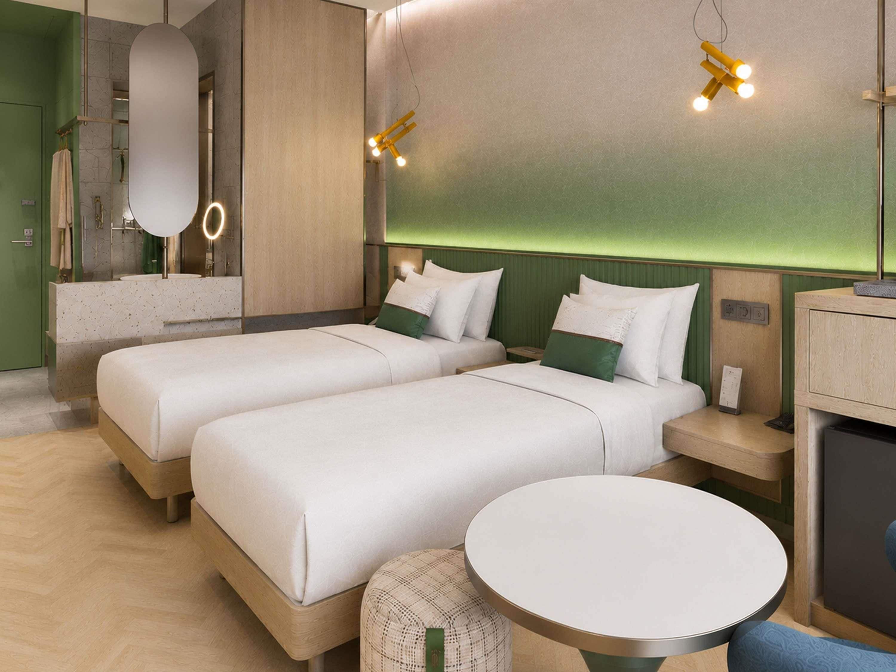 A hotel room with two beds, a round table, a mirror, and a green accent wall.