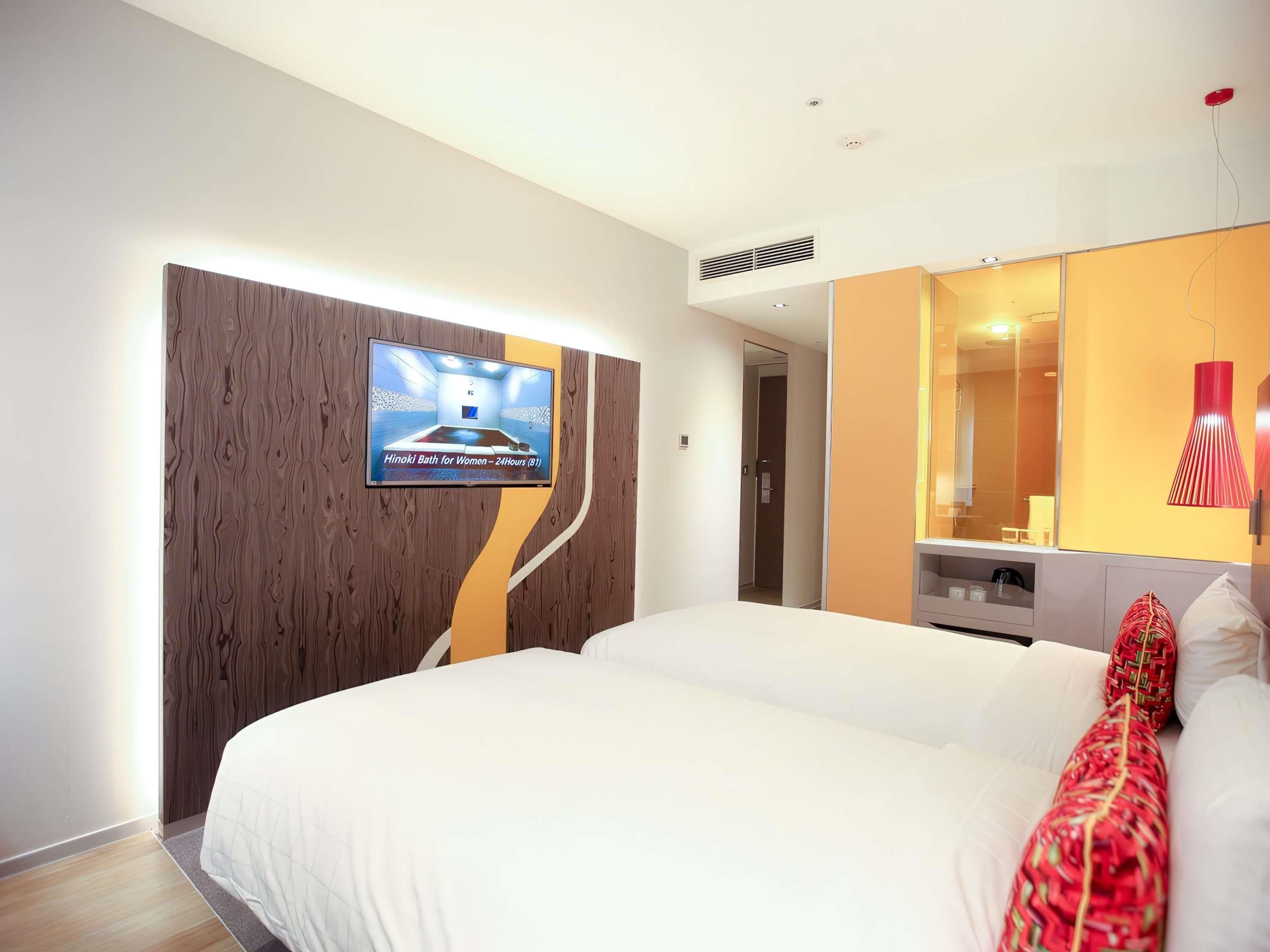 A hotel room with a large bed, a television mounted on a wooden panel, a mirror, and a red pendant light.