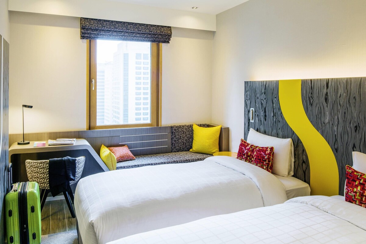 Ein Hotelzimmer mit zwei Betten, einem Schreibtisch und einem Fenster mit Blick auf Gebäude.