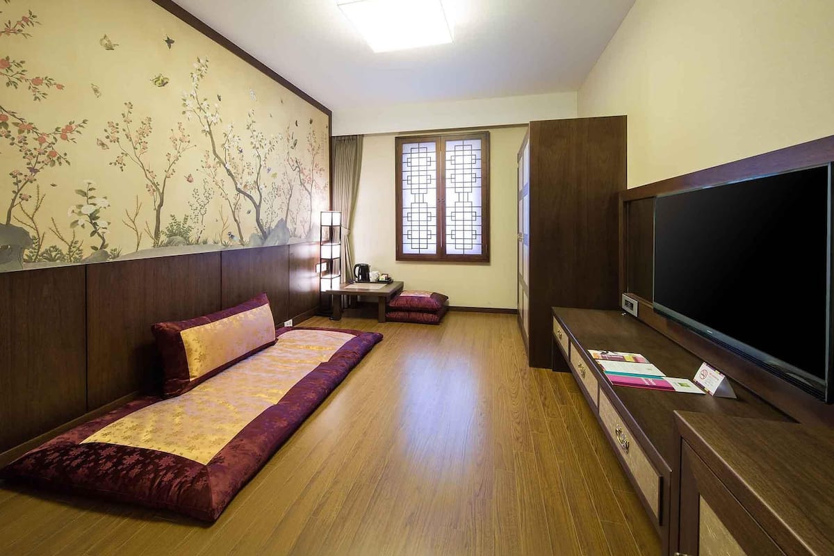 Une chambre d’hôtel comprenant un lit, une télévision, un bureau avec une lampe, une fenêtre donnant sur un paysage agréable et un mur orné d’une fresque florale.