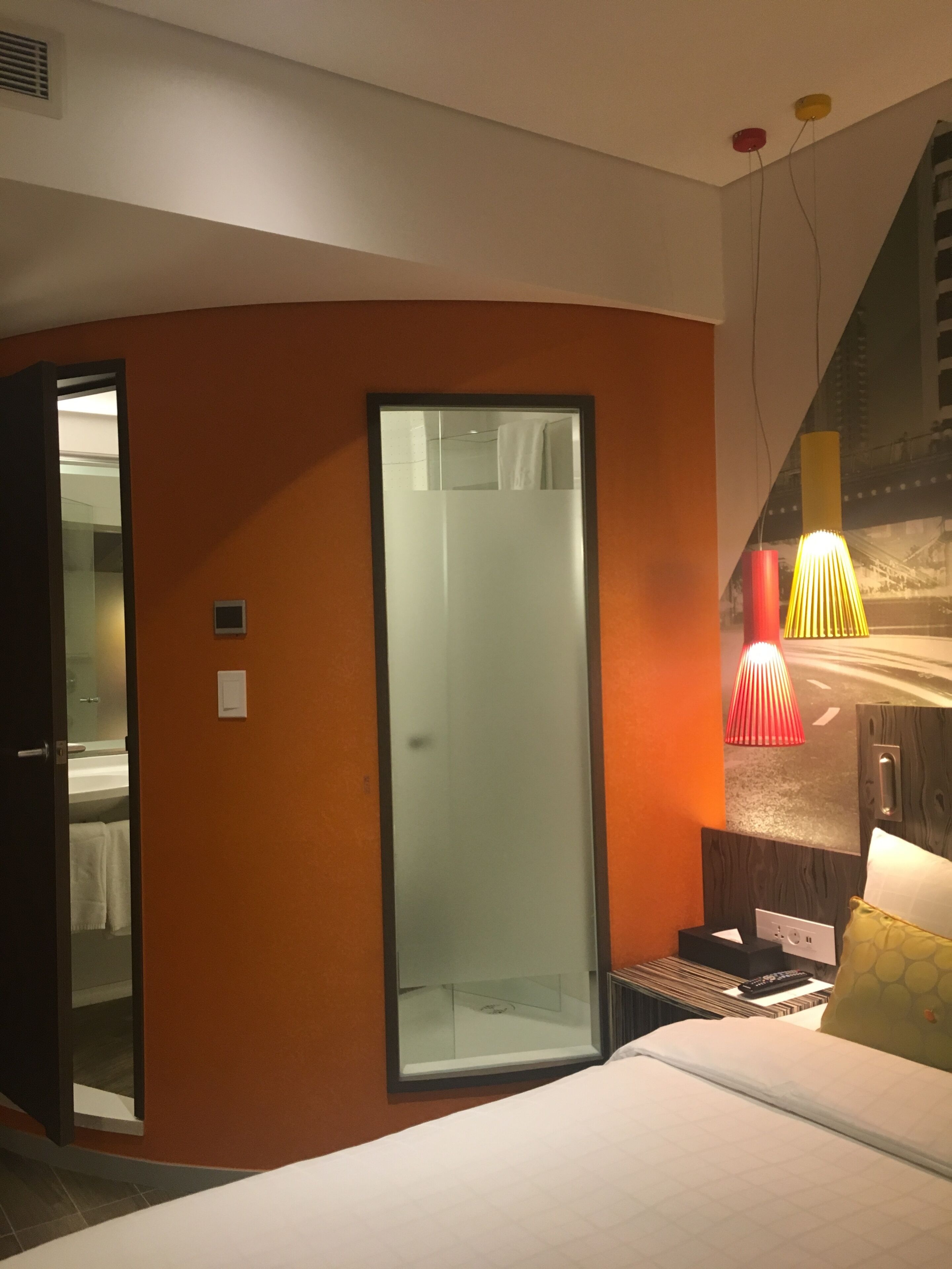 Habitación de hotel con cama, puerta de vidrio que da al baño, una mesita con un control remoto y una lámpara de pared.
