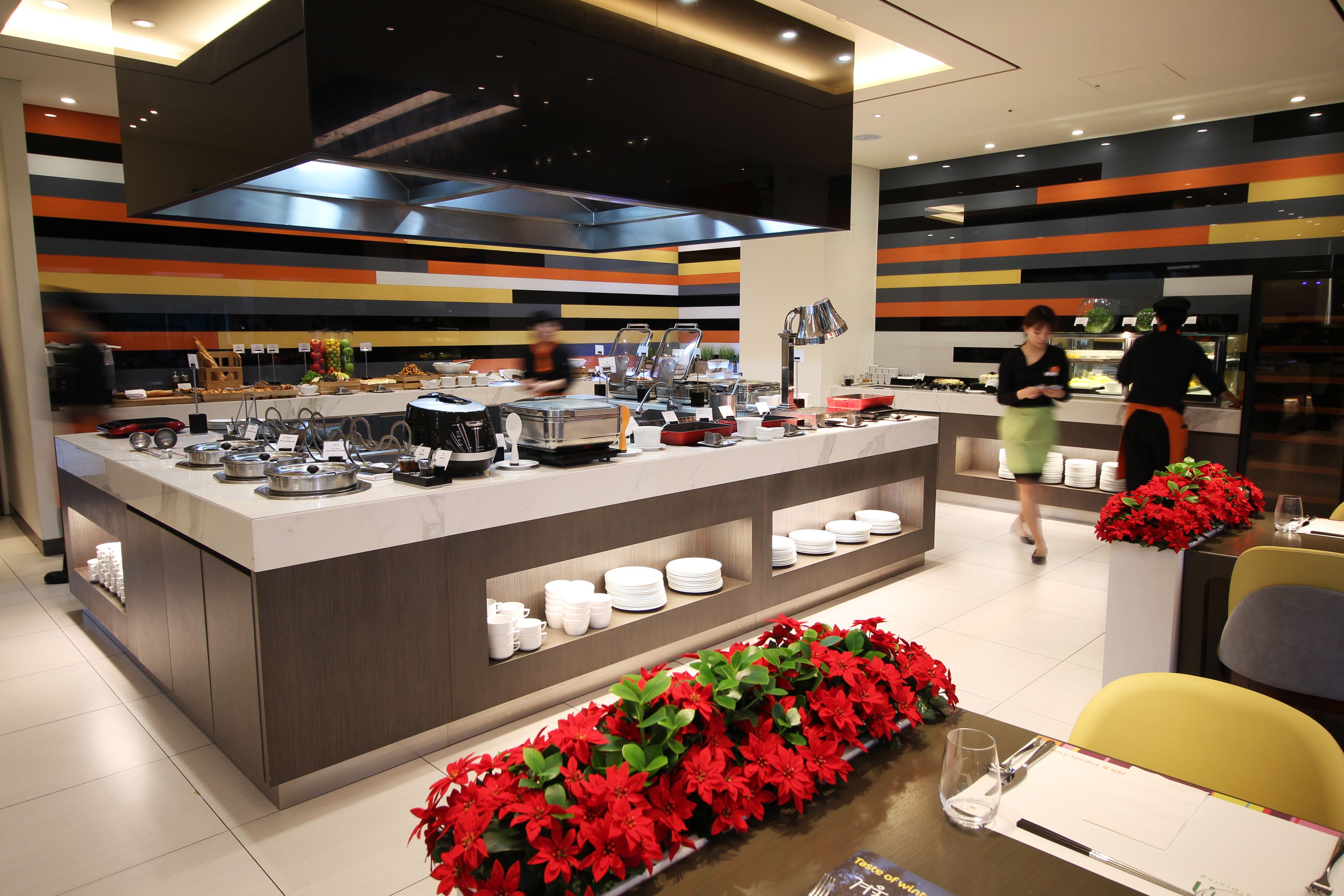 A modern hotel kitchen with a central island, buffet setup, and a dining area with yellow chairs.