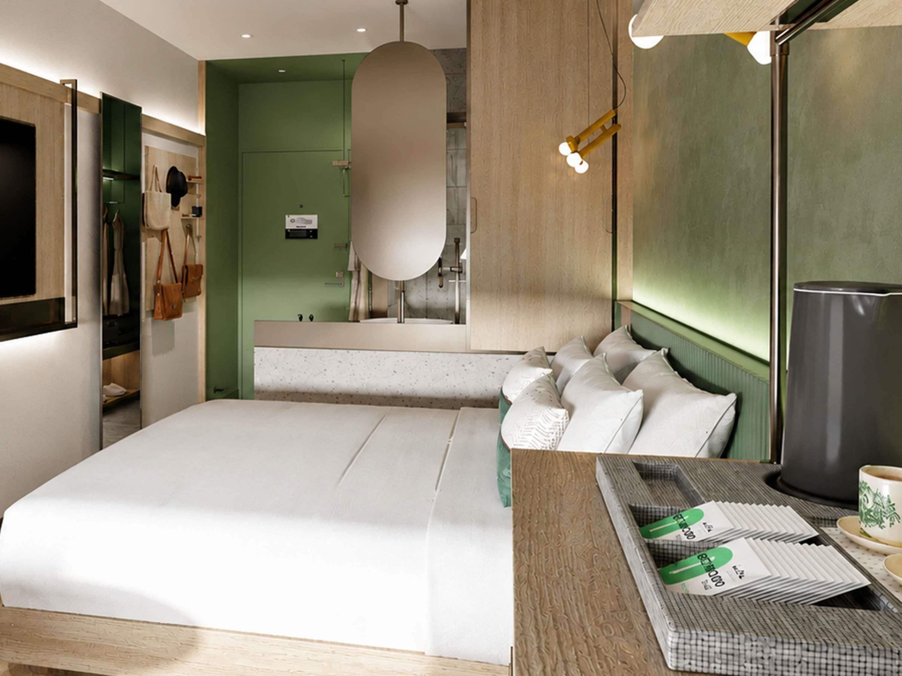 A hotel room with a bed, a bedside table with a cup and papers, a mirror, and a green wall.