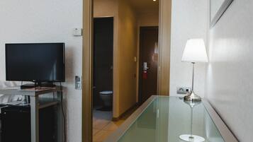 A hotel room with a flat-screen TV on a stand, a glass-top table with a lamp, and a door leading to a bathroom.