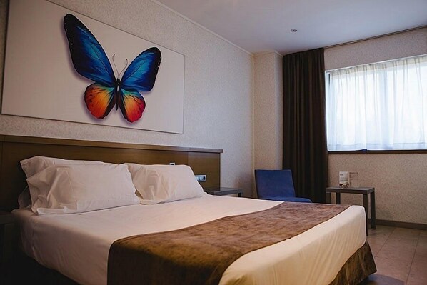 A hotel room with a large bed, a nightstand, a chair, a small table, and a window with curtains. - Hotel Mas Camarena (Paterna)