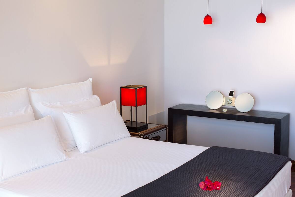 A neatly made bed with white linens and a dark throw blanket, a red lamp, and a bedside table with a clock and a mirror.