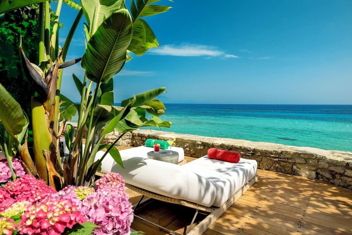 A wooden deck with a white sofa and red pillow, overlooking a turquoise sea.