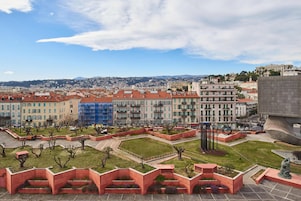 Panorama urbano con un parque que incluye macetas rojas y una estatua, rodeado de edificios con tejados rojos y una vista distante de colinas.
