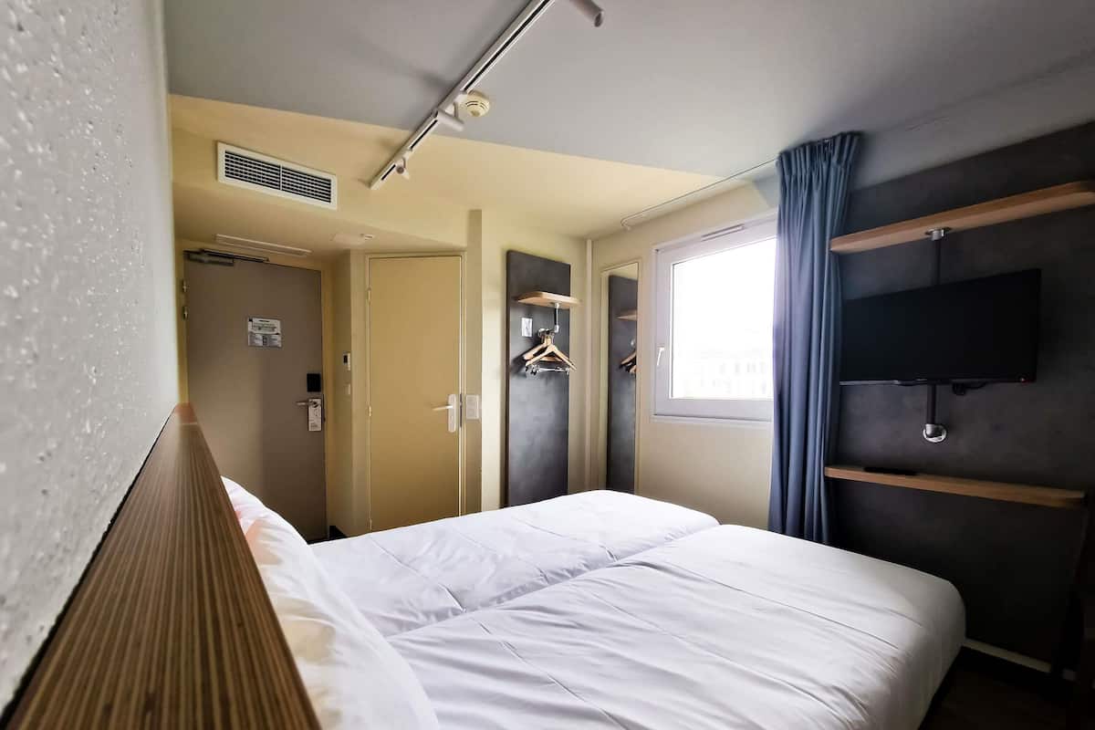 Hotellihuone, jossa on sänky, seinälle asennettu televisio, ikkuna verhoilla, ovi ja ilmanvaihtojärjestelmä.