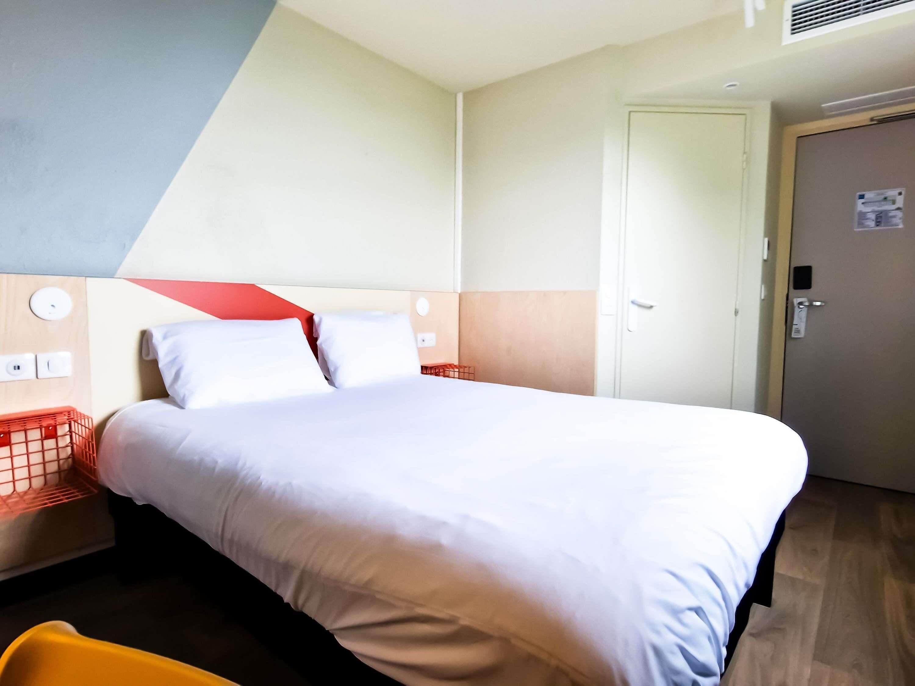 Habitación de hotel con cama, mesita de noche, cesta con marco rojo, puerta y pared con diseño geométrico.