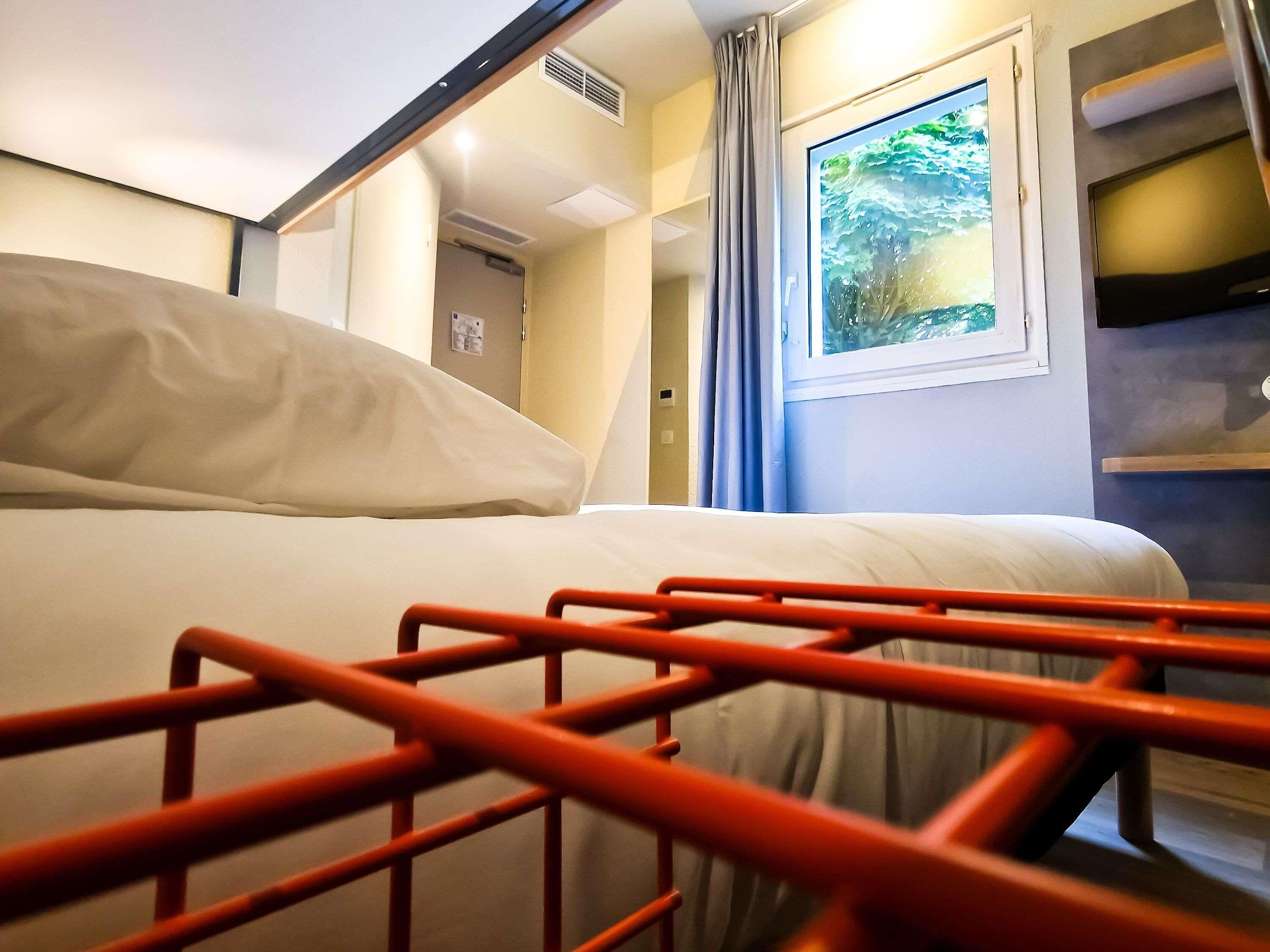 A hotel room with a bed, a window with a view, a door, and a wall-mounted shelf.
