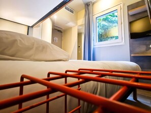 A hotel room with a bed, a window with a view, a door, and a wall-mounted shelf. - ibis budget Lille Marcq-en-Baroeul (Marcq-en-Baroeul)