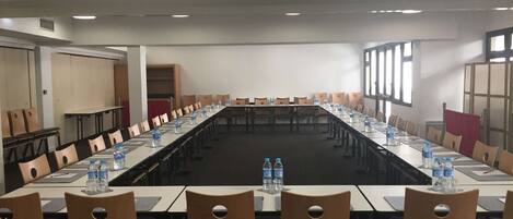 Un salón de conferencias con mesas largas y sillas, botellas de agua y vistas al exterior a través de grandes ventanales.