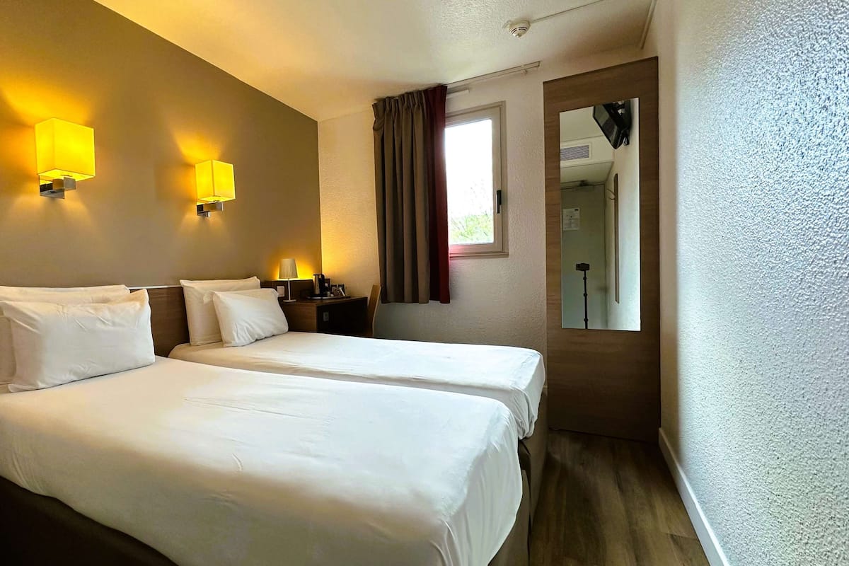 A hotel room with two beds, a nightstand, a lamp, a window with curtains, and a mirror.