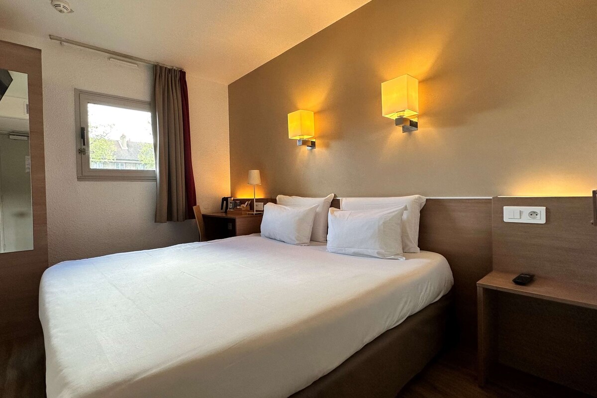 Une chambre d’hôtel avec un lit bien fait, une table de chevet avec une lampe, un miroir et une fenêtre avec des rideaux.