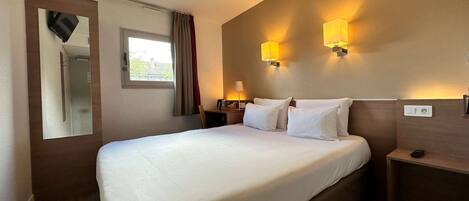 Une chambre d’hôtel avec un lit bien fait, une table de chevet avec une lampe, un miroir et une fenêtre avec des rideaux.