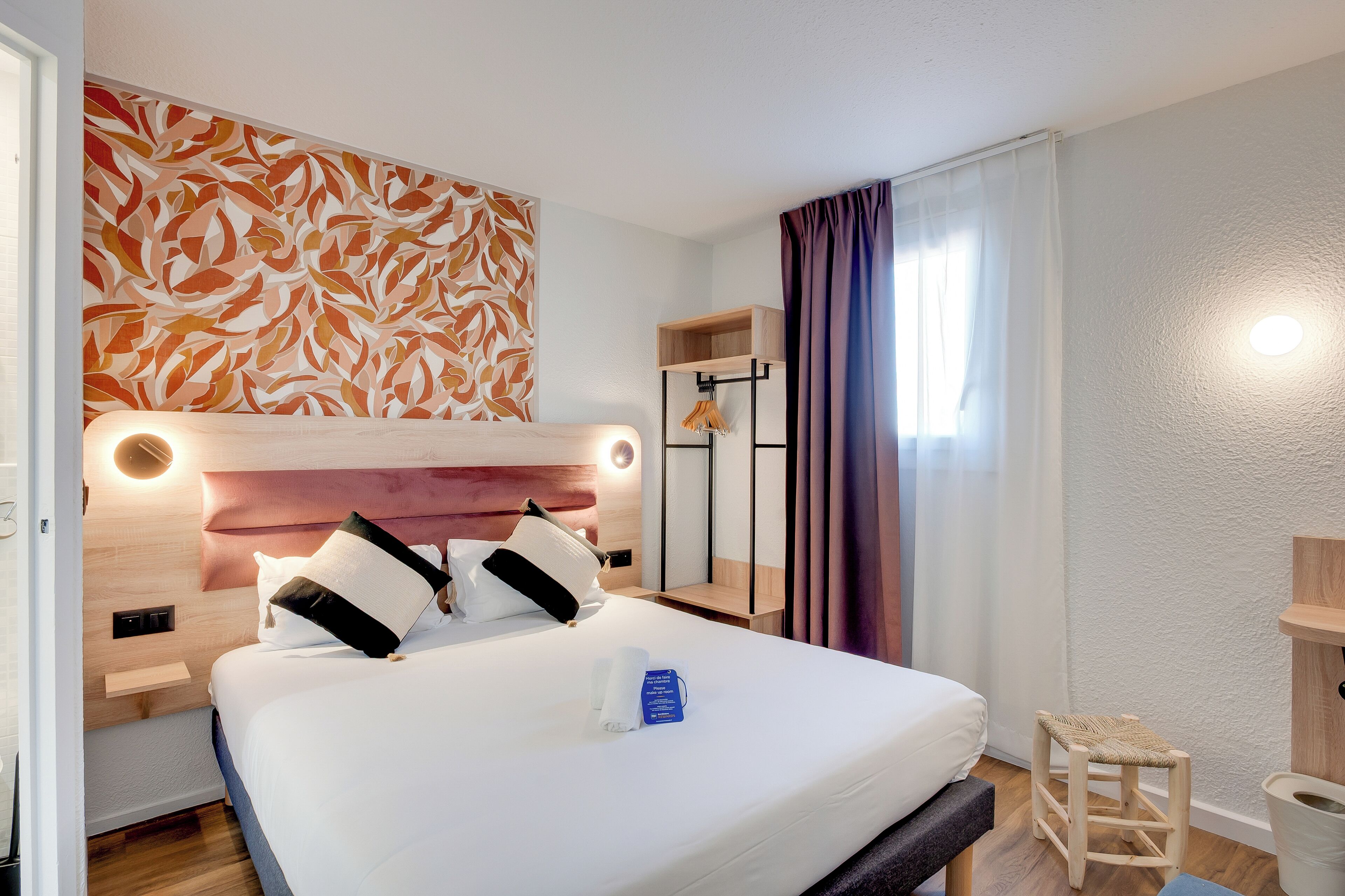 Una habitación de hotel moderna con una cama grande, un cabecero con iluminación integrada, una mesita de madera, un taburete pequeño y una estantería en la pared.