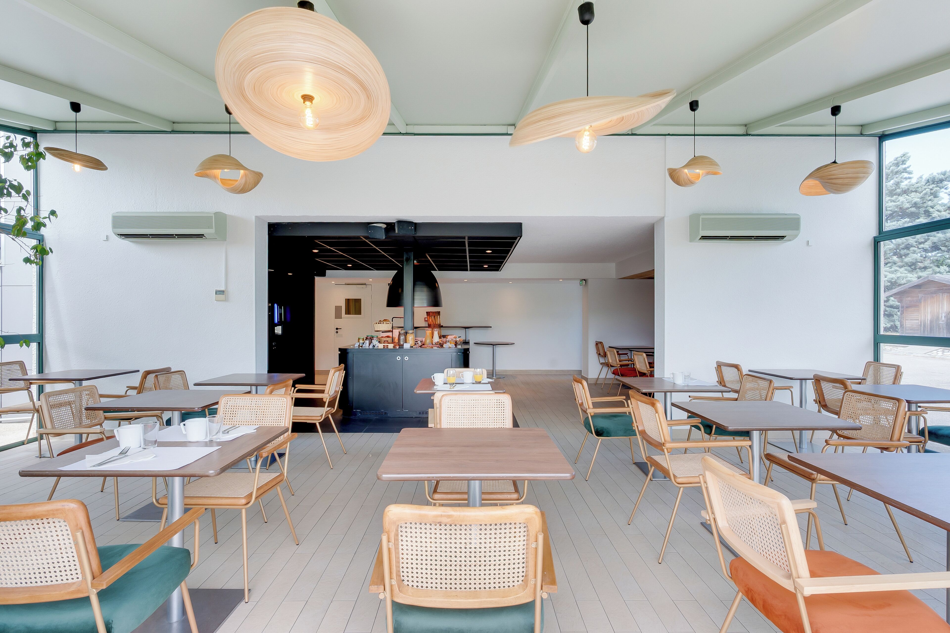 Un comedor amplio con mesas y sillas de madera, iluminación colgante y vistas a la cocina al fondo.