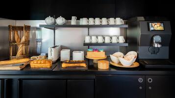 Montaje de desayuno buffet con pan, pasteles, máquinas de café y diferentes recipientes.