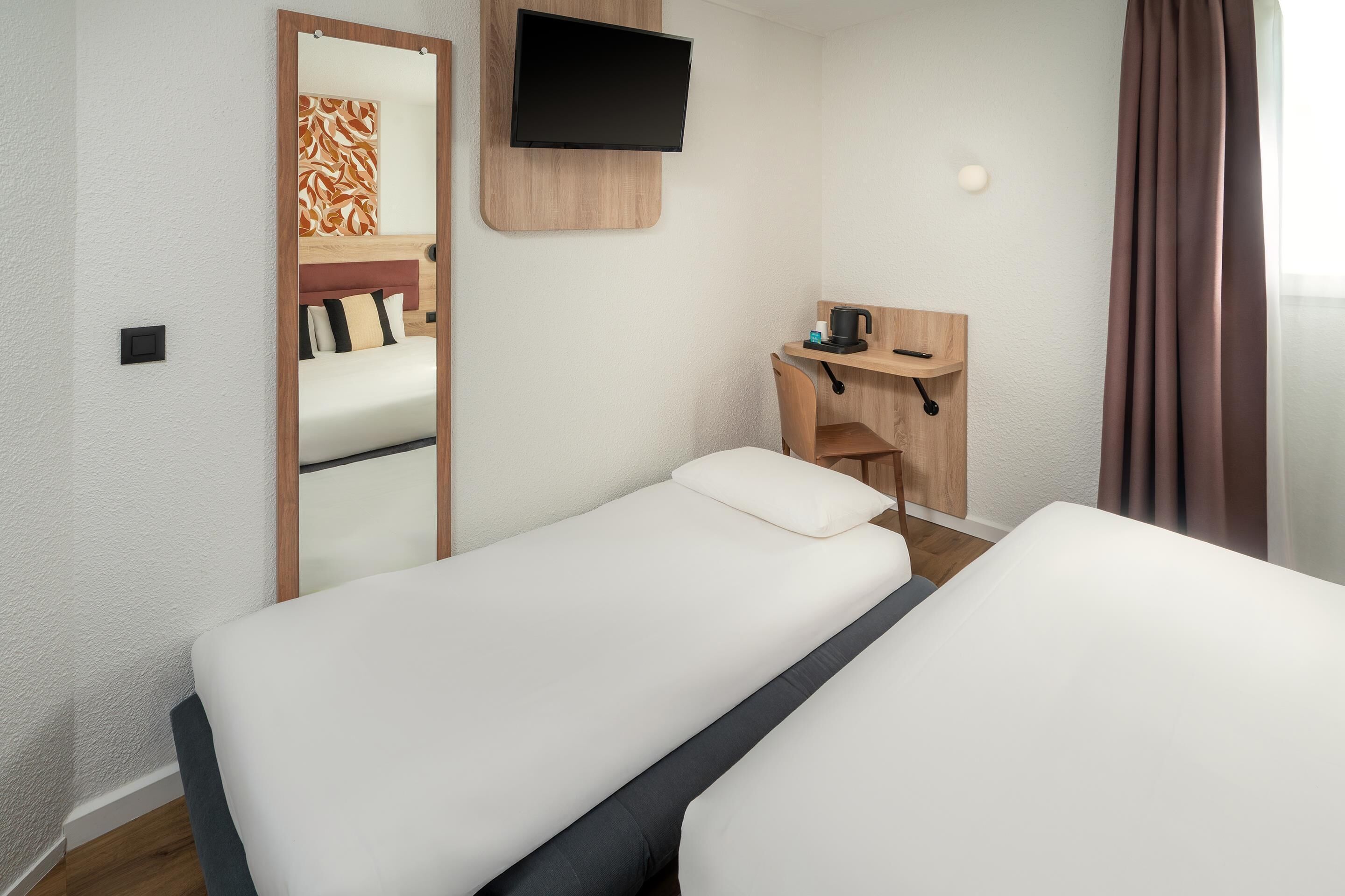 Une chambre d’hôtel avec deux lits simples, un bureau avec une chaise, un téléviseur fixé au mur et un petit coin salon avec une table et des chaises.