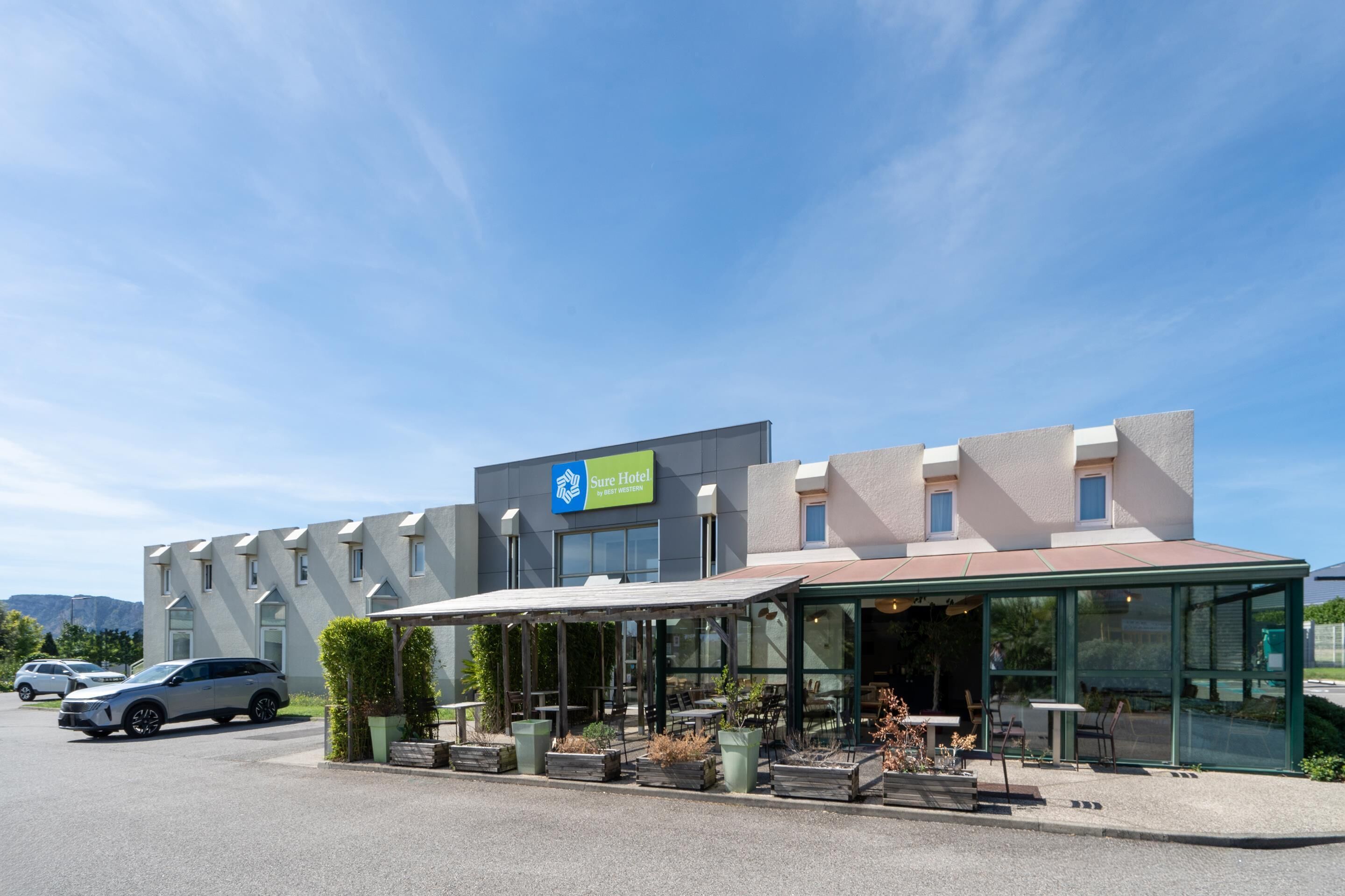 Un hotel moderno con un área de estar cubierta al aire libre, un estacionamiento y vistas a la montaña al fondo.