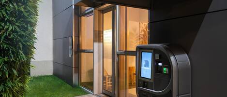 Una entrada de edificio moderna con un teclado digital para acceso, puertas de cristal y un pequeño área de estar al aire libre visible a través de la entrada.