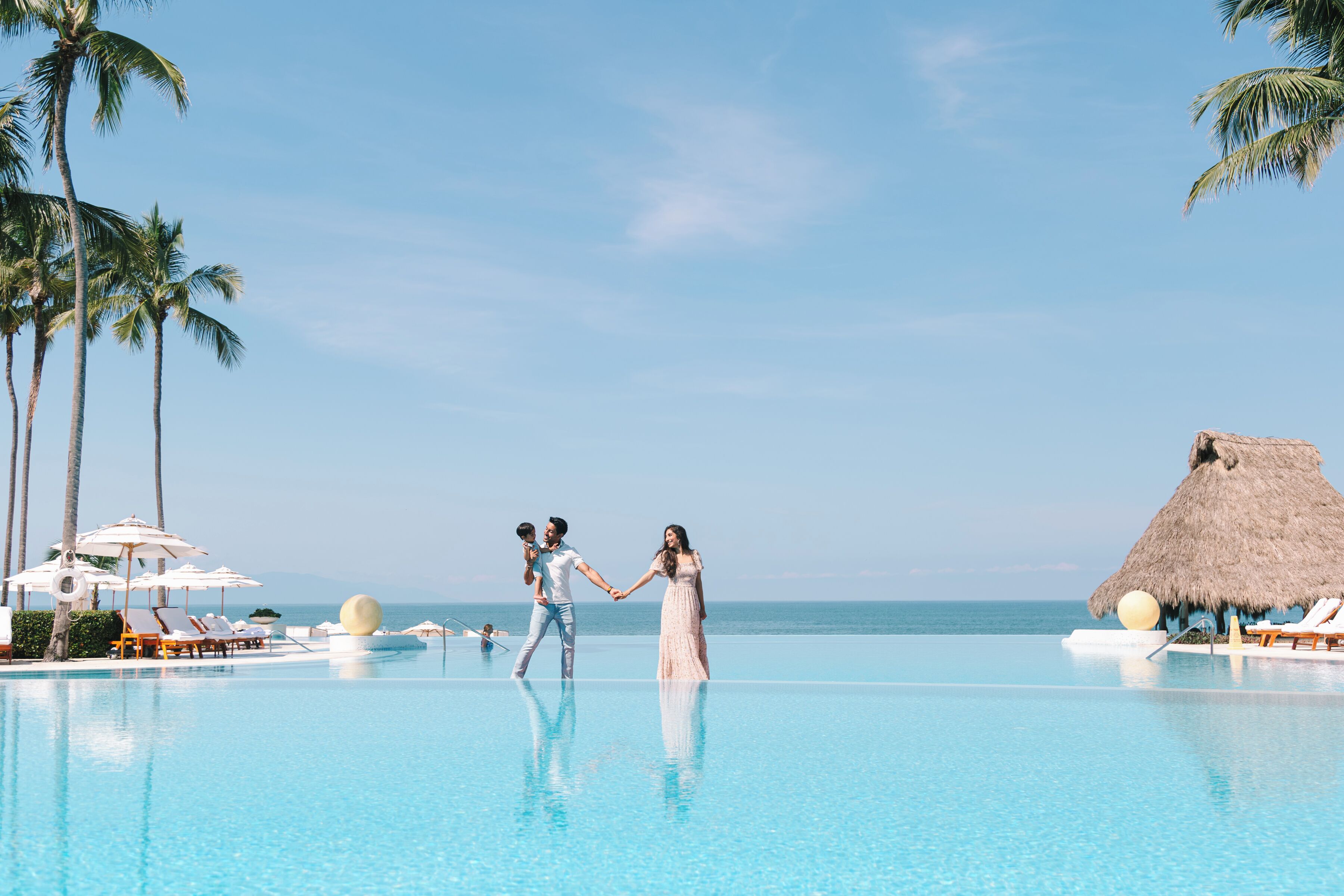 A couple holding hands in a pool with lounge chairs and palm trees, overlooking the ocean.